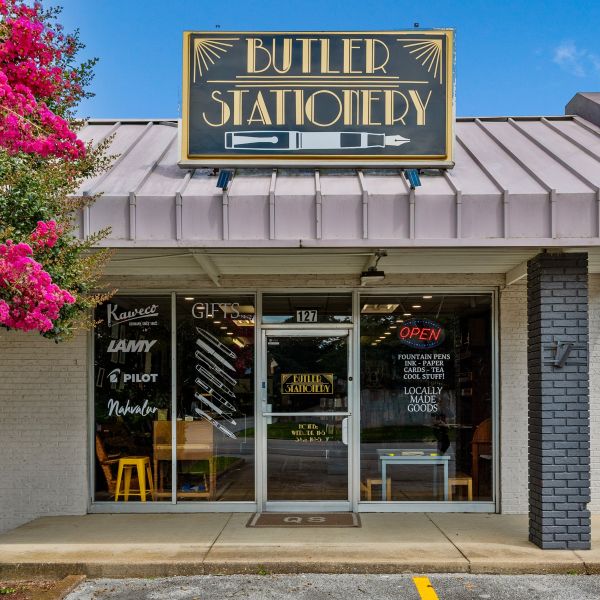 Front door and windows of Butler Stationery shop with large art deco sign with Butler Stationery text and fountain pen graphic on the roof and parking spaces and flowering tree out front.