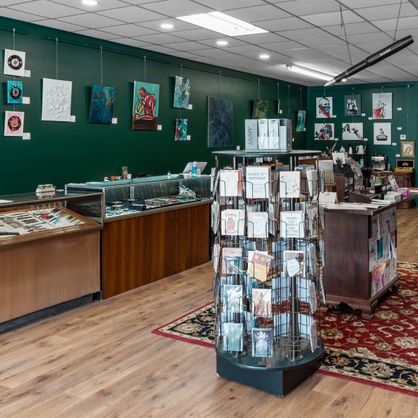 View inside the Butler Stationery shop to the left, with dark green walls and hanging art behind display cases of pens, then a greeting card spinner and island table displaying more merchandise in the center of the room.