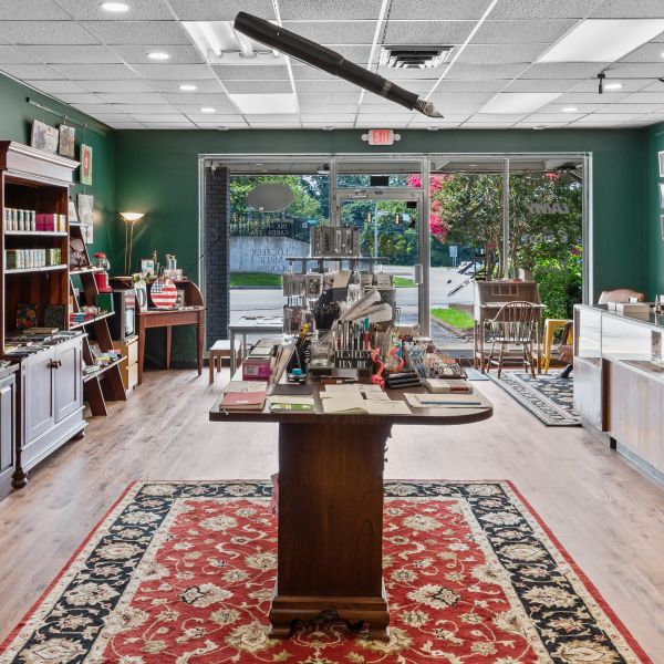 View from the back of the Butler Stationery shop toward the front windows showing the center island of pens, pencils, paper, and other stationery items, with a large Kaweco sport model hanging from the ceiling above, glass pen display cases along the right, and wooden shelves with more stationery, paper, mugs, tea, leather items, etc. along the left.