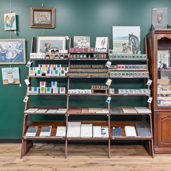 View inside the Butler Stationery shop of the dark green side wall with hanging art beside and above wooden shelves with bottles of fountain pen ink, paper, refills, 3D printed accessories, mugs, notebooks, etc.