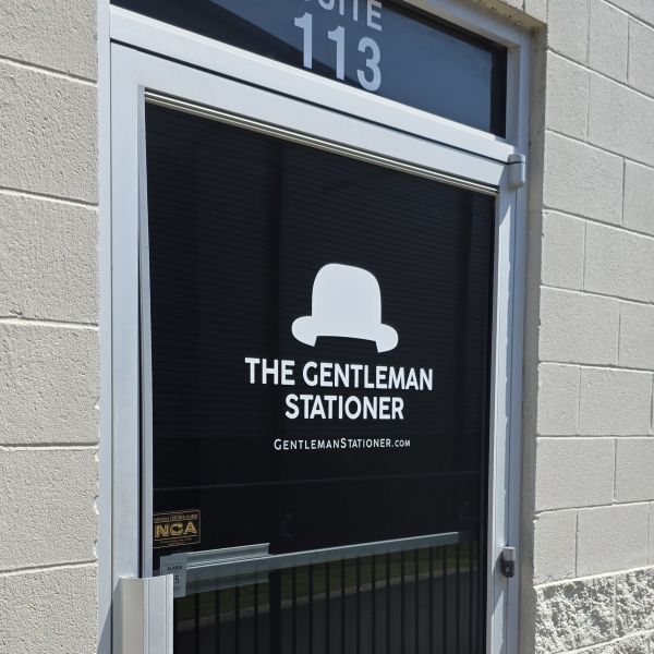 Collage of two photos. View of the walkway to The Gentleman Stationer shop on a bright sunny day, with standing 'OPEN' sign guiding visitors to the door.Front glass door of The Gentleman Stationer shop with logo and website address on the door, Suite 113.