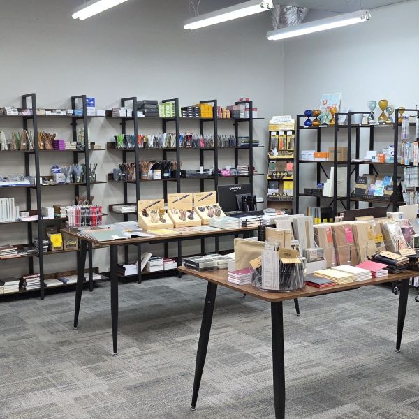 View inside The Gentleman Stationer shop with a spinner of Lamy pens and pencils, multiple shelves along the walls with pencils, paper, notebooks, planners, multipens, sand glasses, highlighters, etc., and two tables with multiple notebook systems including Roterfaden Taschenbegleiter, Lochby, and Travelers, and other writing tools and stationery.