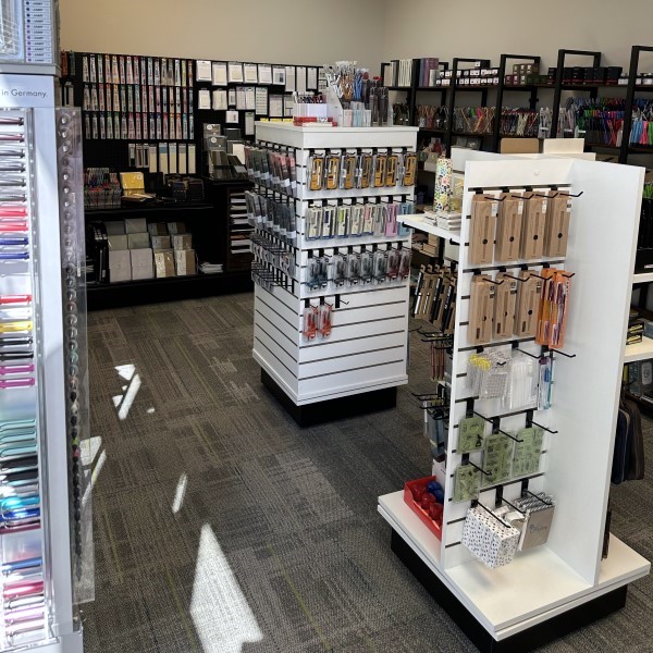 View inside The Gentleman Stationer shop with a spinner of Lamy pens and pencils, multiple shelves along the walls with pencils, paper, notebooks, planners, multipens, highlighters, etc. and two freestanding spinners with dip pens, stamps, fountain pens, gel pens, ballpoint pens, sharpeners, and other writing tools and stationery.